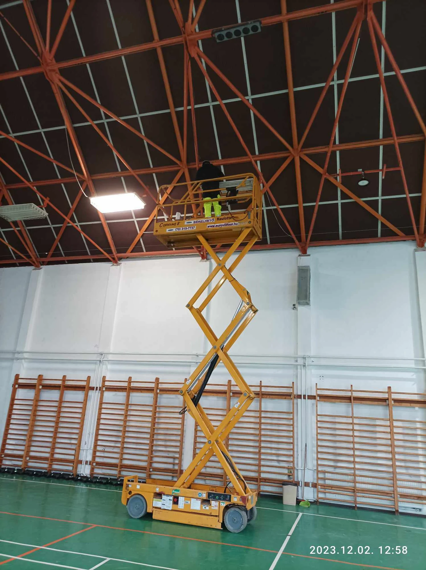 Szakember sárga ollós emelőről dolgozik a sportcsarnok mennyezeténél.